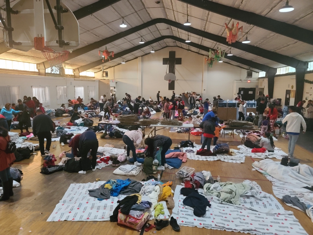 Dozens of migrants of all ages gather in the gym near Sacred Heart parish in El Paso, Texas where they are placing sheets to sleep on the wooden floor of the gym. Visible is a large wooden cross behind a basketball backboard and hoop.